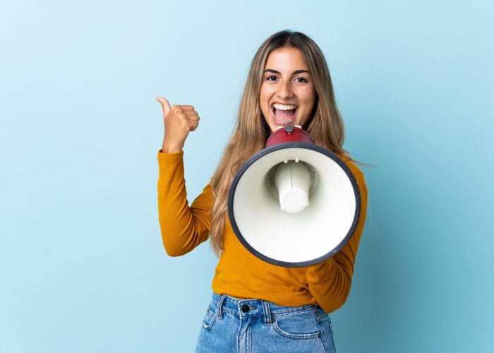 Young hispanic woman over isolated blue background shouting through a megaphone and pointing side